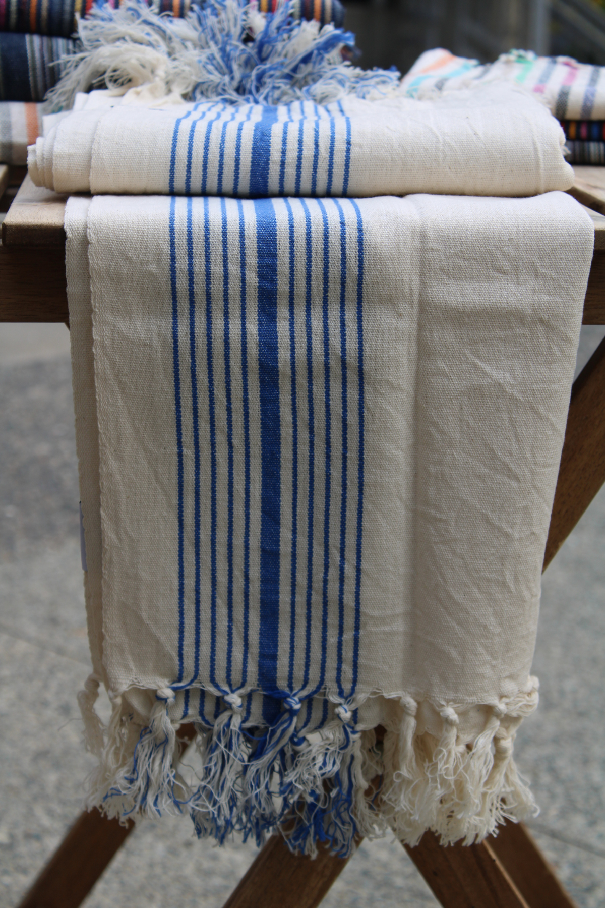 Experience the beauty of our Organic Turkish Hand Towel, elegantly draped from the table for a close-up view. Featuring exquisite tassels and a high-quality design in white with blue stripes, this towel is available at Soumak Boutique, Vancouver, BC, Canada, inside the Kids Market on Granville Island.